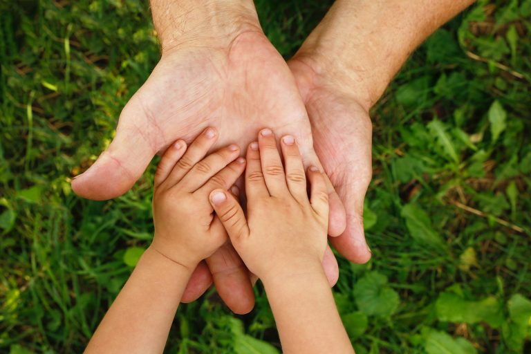 the small hands of a child resting on the hands of an adult with grass in the background
